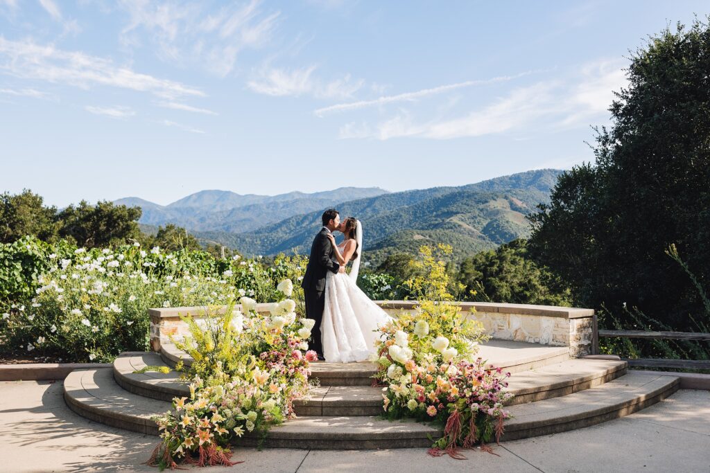Bride and groom kissing at Holman Ranch ceremony terrace with panoramic Carmel Valley mountain views and lush floral arrangements in peach, blush, and yellow.