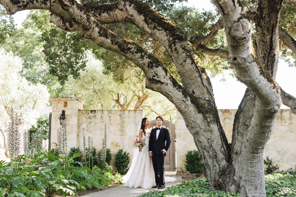 Bride and groom wedding portrait in front of historic stone building at Holman Ranch, Carmel Valley, bride holding romantic floral bouquet.